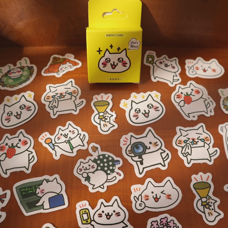 May include: A collection of cat-themed stickers and a yellow box. The stickers feature various cartoon cats with different expressions and holding objects like microphones and strawberries. The box reads "NIZMO-CARD" and "Cat's Mood".