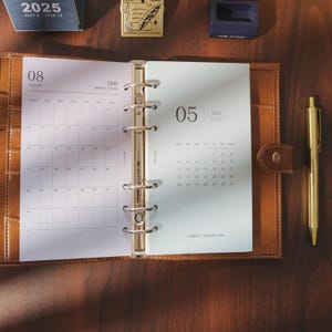 May include: Open brown leather planner with gold accents, displaying calendar pages for August and May 2026. A gold pen rests beside the planner. The planner is open on a dark wooden surface.