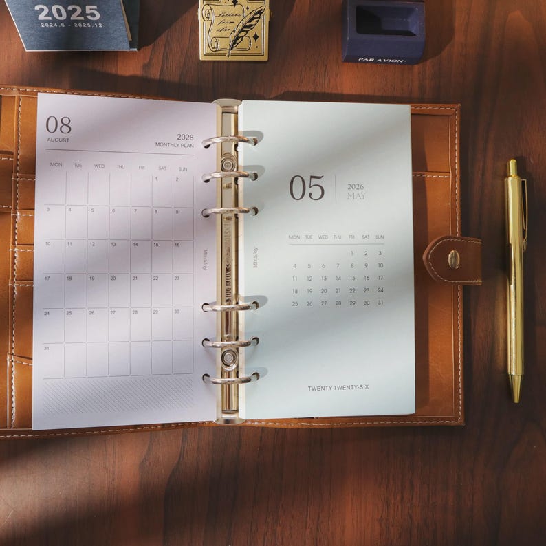 May include: Open brown leather planner with gold accents, displaying calendar pages for August and May 2026. A gold pen rests beside the planner. The planner is open on a dark wooden surface.