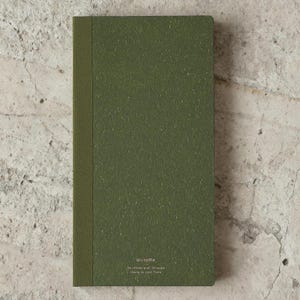 May include: A closed, olive green notebook with a textured cover and a darker spine. The notebook has the text "WriteMe" and other details printed at the bottom. The notebook is rectangular and appears to be new.