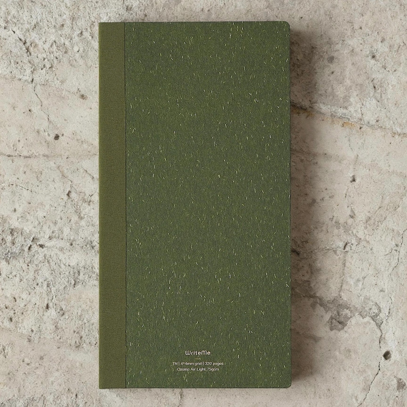 May include: A closed, olive green notebook with a textured cover and a darker spine. The notebook has the text "WriteMe" and other details printed at the bottom. The notebook is rectangular and appears to be new.