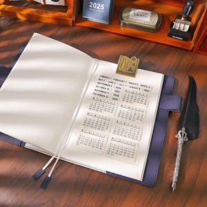 May include: A blue leather journal with a calendar insert, open to display the dates for 2025-2029. A gold-colored book-shaped paper clip and a decorative quill pen are also visible. The journal has a ribbon bookmark.