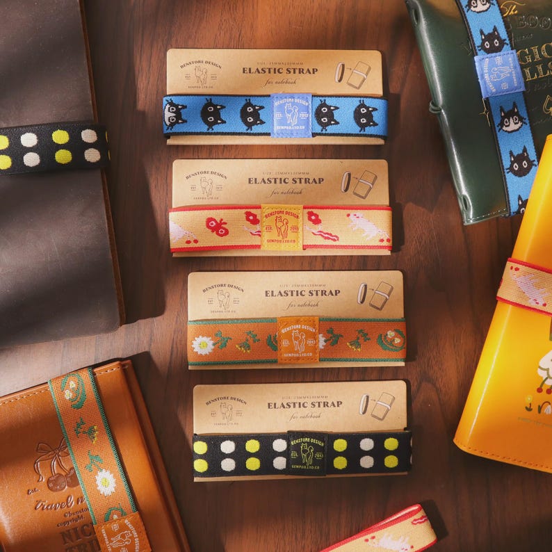 May include: Notebook elastic straps in various designs. The straps feature patterns including black cat illustrations, floral motifs, and polka dots. Each strap is packaged with a cardboard label that reads "Elastic Strap for notebook."