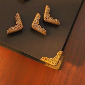 Puede incluir: Protectores de esquina de latón ornamentados, de estilo antiguo. Estas piezas metálicas decorativas presentan un diseño en espiral y están colocadas sobre una superficie oscura. Probablemente se utilizan para proteger las esquinas de una caja, un libro u otro objeto.