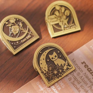 Może przedstawiać: Trzy dekoracyjne metalowe klipsy w kolorze mosiądzu. Jeden klips przedstawia kota na księżycu w kształcie półksiężyca z gwiazdami i kwiatami. Inny klips ma wzór róży i księżyca. Trzeci klips ma kwiatowy wzór.