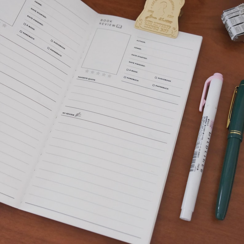 May include: Open notebook with lined pages for book reviews, featuring sections for author, genre, and date. A white and pink highlighter and a green pen rest on the wooden surface. A decorative bookmark is at the top of the notebook.
