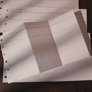 May include: A collection of white planner pages, each marked with "MONTHLY PLANS". The pages are folded open, displaying a calendar grid with numbered days and space for notes. The pages are pre-punched for binding.