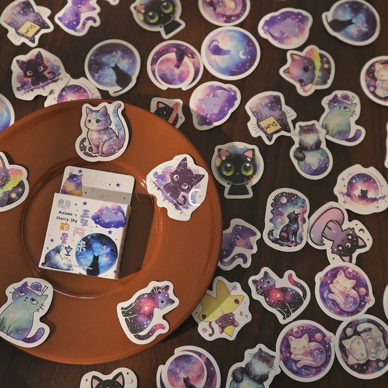 May include: A collection of cat-themed stickers in various designs, including cats with galaxy patterns, moons, and stars. The stickers are in shades of purple, blue, and white. A small box of stickers is also visible, with text in a foreign language.
