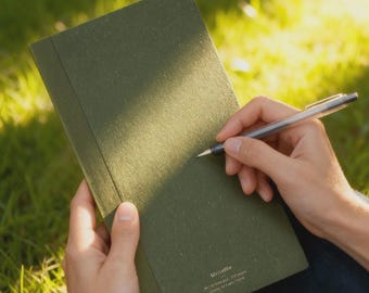 Può includere: Un taccuino verde oliva aperto, con una penna pronta per scrivere. Il taccuino ha una copertina strutturata e il testo "WriteMe" è visibile. La penna è argento e nera. Lo sfondo è erba verde sfocata.
