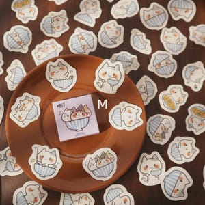 May include: A collection of cat-themed stickers featuring various poses and activities. The stickers are primarily white with gray accents, set against a light blue striped background. A small notepad with a cat illustration is also present.