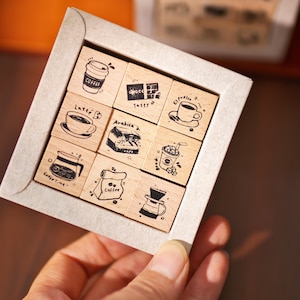May include: A set of nine wooden stamps featuring coffee-related illustrations and text, such as coffee cups, beans, and brewing equipment. The stamps are arranged in a square within a grey cardboard box, held by a hand.