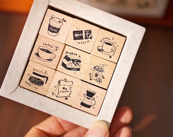 May include: A set of nine wooden stamps with coffee-themed illustrations and text, including coffee cups, beans, and brewing equipment. The stamps are arranged in a square within a gray cardboard box, held by a hand.