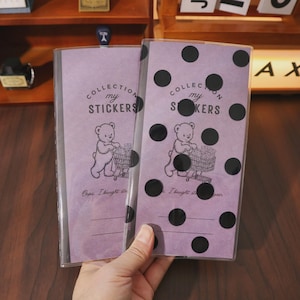 May include: Two clear sticker books with purple covers. The covers have black polka dots and a cartoon bear pushing a shopping cart. The text "COLLECTION my STICKERS" and "Oops. I bought stickers again" are printed on the covers.