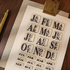 Könnte beinhalten: Ein Set aus transparenten Acrylstempeln mit den Monatsnamen in fetter, schwarzer Schrift. Das Set enthält einzelne Stempel für jeden Monat sowie einen dekorativen Stempel in Buchform. Ein goldener Stift liegt auf der Holzoberfläche.