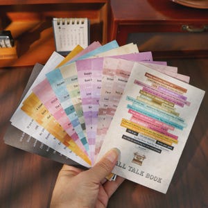 Può includere: Un ventaglio di cartoncini rettangolari con varie citazioni ispiratrici stampate su sfondi colorati. La carta superiore mostra il testo ALL TALK BOOK sopra una piccola illustrazione di una macchina da scrivere.