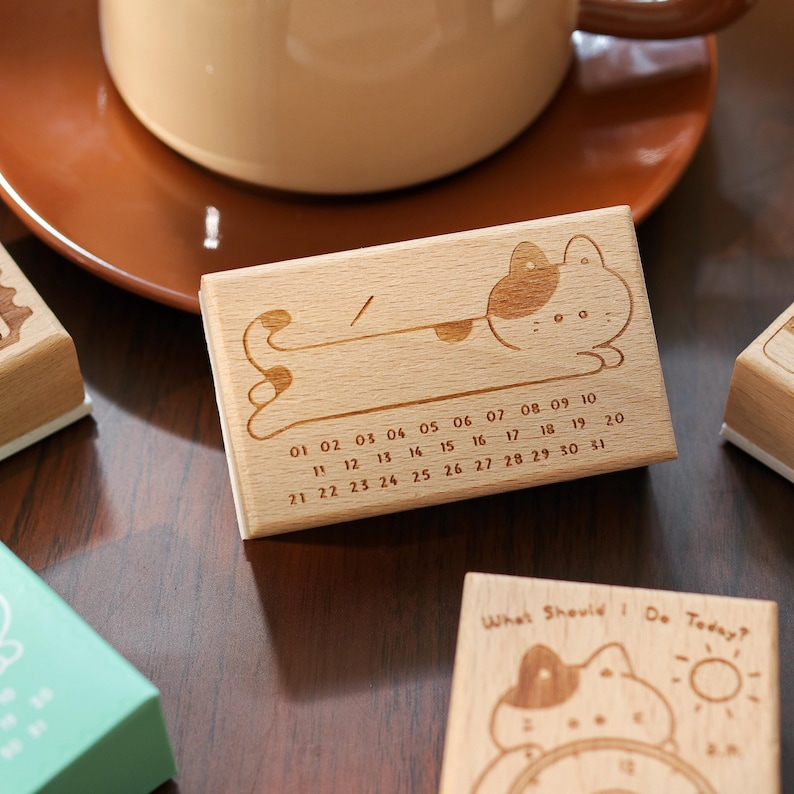 May include: A wooden calendar stamp with a cat illustration and the numbers 01-31. The stamp is rectangular and made of light-colored wood. The cat is lying down with a dotted pattern. The text "What Should I Do Today?" is on a separate stamp.