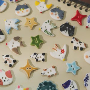 Puede incluir: Una colección de botones de cerámica en varias formas y colores. Los botones presentan diseños como gatos, pájaros, estrellas y perros. Los botones son blancos, con detalles en azul, amarillo, rojo, verde y marrón. Ideales para proyectos de costura y manualidades.