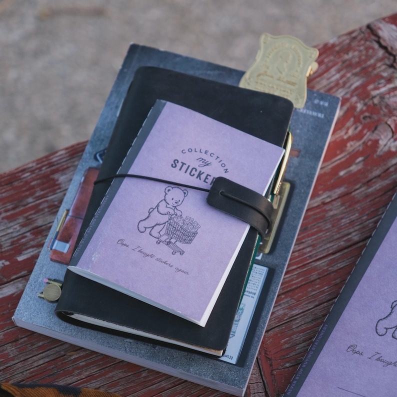 May include: A stack of notebooks including a light purple sticker collection booklet featuring a bear pushing a shopping cart with the text Collection my STICKER and Oops I bought stickers again, secured by a black elastic band on a black cover.