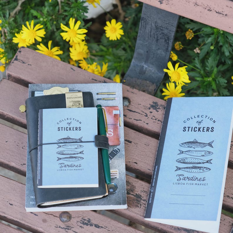 May include: A collection of notebooks and stickers with a Lisbon Fish Market theme. The notebooks are black and light blue, with the text "COLLECTION OF STICKERS" and "Sardines" printed on them. A green pen is attached to the notebook.
