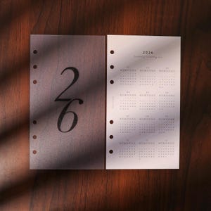 May include: Two planner inserts with the numbers "26" and the year "2026" printed on them. The "26" is in a large, elegant font on a semi-transparent sheet. The other sheet displays a calendar grid with the year 2026.