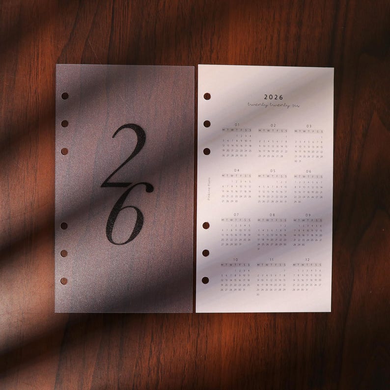 May include: Two planner inserts with the numbers "26" and the year "2026" printed on them. The "26" is in a large, elegant font on a semi-transparent sheet. The other sheet displays a calendar grid with the year 2026.