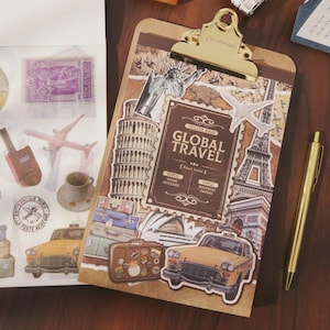 Pode incluir: Uma prancheta com uma presilha dourada segurando um livro de adesivos "Global Travel". O livro de adesivos apresenta imagens de marcos, aviões e bagagens. Uma caneta dourada repousa sobre a superfície de madeira. Adesivos adicionais com selos e imagens temáticas de viagens são visíveis.