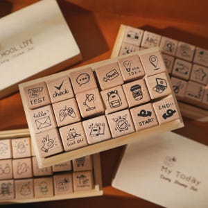 May include: A set of wooden rubber stamps arranged in a rectangular box. Each stamp features a unique black ink icon or text, including symbols like a lightbulb, backpack, alarm clock, game controller, and text such as TEST, START, and END!!.