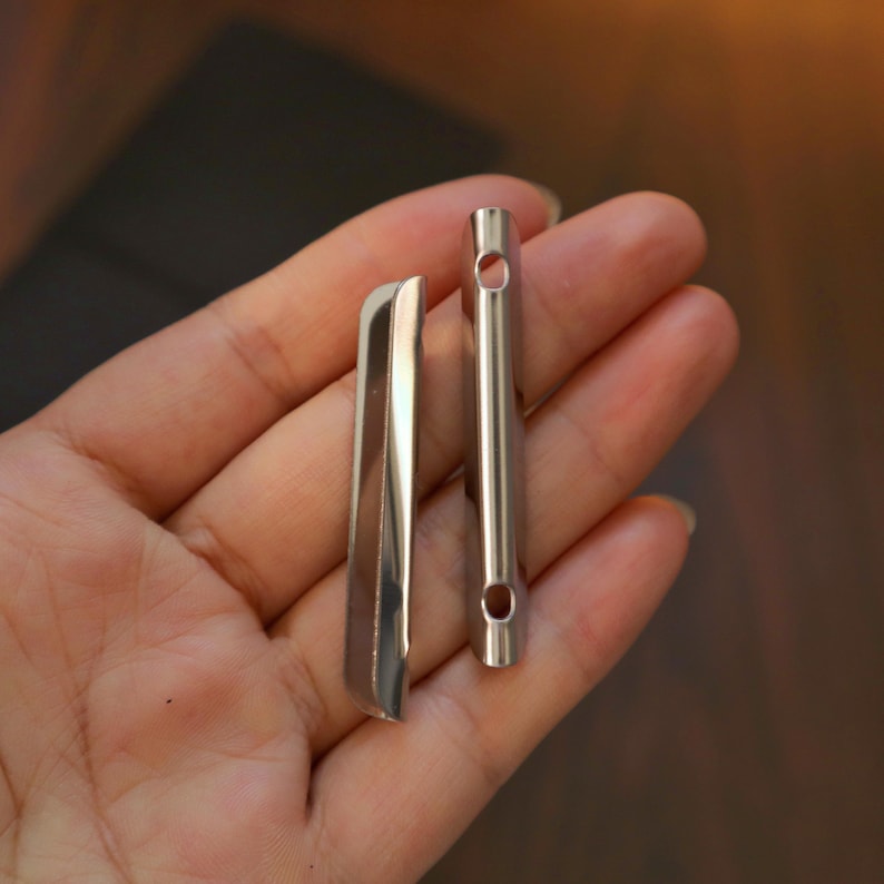May include: Two small, shiny, silver metal objects. One is a slender, rectangular shape with a curved edge. The other is a thin, cylindrical tube with a hole at each end. The objects are held in a hand.