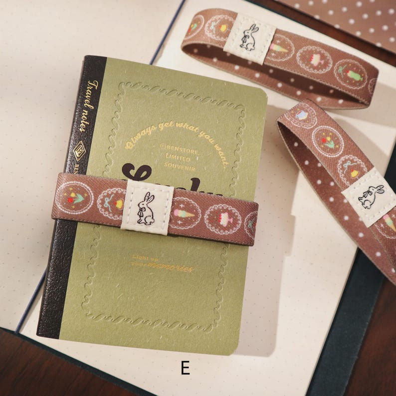 May include: A green notebook with a brown elastic band featuring a white bunny and decorative patterns. The notebook has the words "Travel notes" and "Always get what you want" printed on it. Additional bands are visible.
