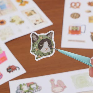 May include: A die-cut sticker featuring a black and white cat wearing a green headscarf patterned with small orange and white floral details. The sticker is held by light blue metal tweezers, showcasing the detailed illustration of the cat's face.