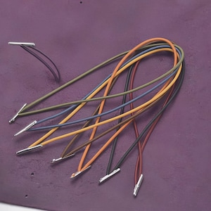 May include: A collection of colorful elastic cords with silver metal tips, arranged on a textured purple surface. The cords are in various colors, including orange, blue, green, and brown. The cords are likely for crafting or jewelry making.