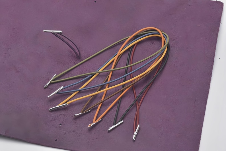 May include: A collection of colorful elastic cords with silver metal tips, arranged on a textured purple surface. The cords are in various colors, including orange, blue, green, and brown. The cords are likely for crafting or jewelry making.