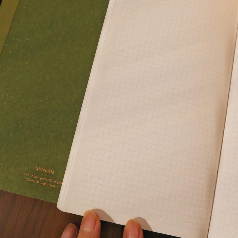 May include: Open notebook with grid paper and a green cover. The notebook is open to a page with a light beige grid pattern. The cover has the text "writeme" and other text below it.