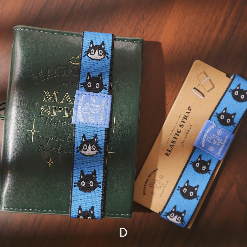 May include: A blue elastic strap with a repeating pattern of black cat faces, used as a bookmark or notebook band. The strap is wrapped around a green notebook and a tan card with the text "ELASTIC STRAP for notebook".