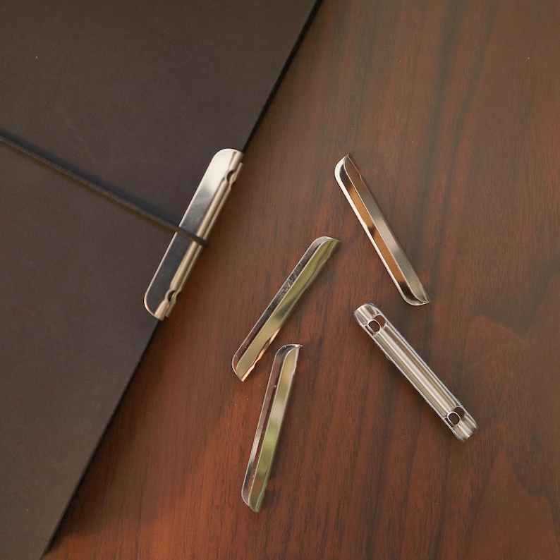 May include: A collection of silver-colored metal binder clips scattered on a wooden surface. The clips are rectangular with rounded edges and a small hole on one end. One clip is attached to a black object.