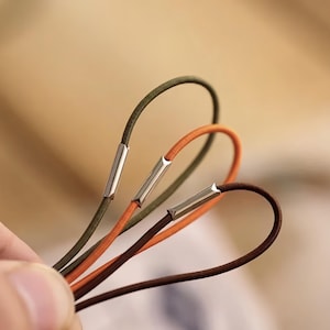 Pu&ograve; includere: Primo piano di tre elastici per capelli verde oliva, arancione e marrone. Ogni elastico ha una chiusura in metallo argentato. Gli elastici sono ad anello e tenuti in mano, con uno sfondo sfocato.