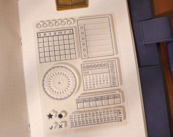 May include: Clear acrylic stamps for bullet journaling. Includes calendar, checklist, and time tracker designs. The stamps are designed to fit in a notebook. The image also shows a gold-colored paper clip.