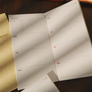 May include: Open weekly planner with a tan cover and white interior pages. The planner is open to a two-page spread with sections for each day of the week, labeled Monday through Sunday. The cover reads "WEEKLY TRAVEL NOTE".