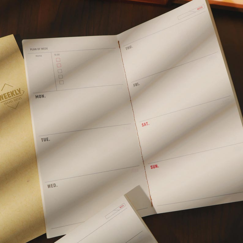 May include: Open weekly planner with a tan cover and white interior pages. The planner is open to a two-page spread with sections for each day of the week, labeled Monday through Sunday. The cover reads "WEEKLY TRAVEL NOTE".