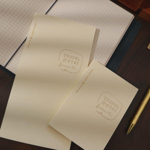 May include: Two cream-colored notebooks with the text "TRAVEL NOTES freewriter" in a gold color. One notebook is open, revealing graph paper. A gold pen rests on a dark brown surface. The notebooks are for writing and journaling.