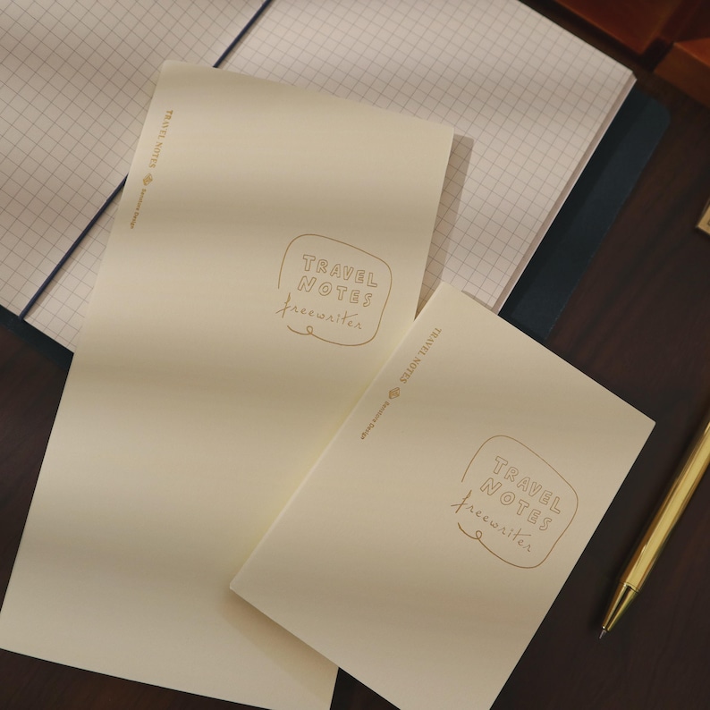 May include: Two cream-colored notebooks with the text "TRAVEL NOTES freewriter" in a gold color. One notebook is open, revealing graph paper. A gold pen rests on a dark brown surface. The notebooks are for writing and journaling.