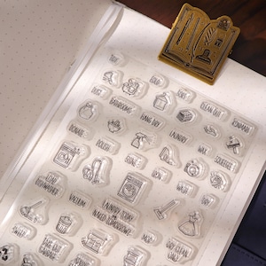 May include: Clear acrylic stamps featuring household chore icons and text, including "Laundry Today or Naked Tomorrow?" and "Dry Clean Only." A gold-coloured metal book-shaped paper clip rests on a dotted notebook page.
