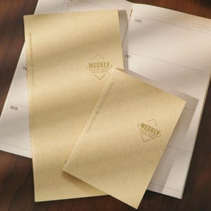 May include: Two beige Traveler's Notebooks with the text "WEEKLY TRAVEL NOTES" in a diamond shape. One notebook is open, revealing a weekly planner layout. The notebooks are on a white and beige surface.