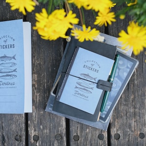 May include: A stack of notebooks and a tablet on a wooden surface, with yellow flowers in the background. The notebooks are light blue with the text "Collection of Stickers" and "Sardines Lisbon Fish Market". A green pen is visible.