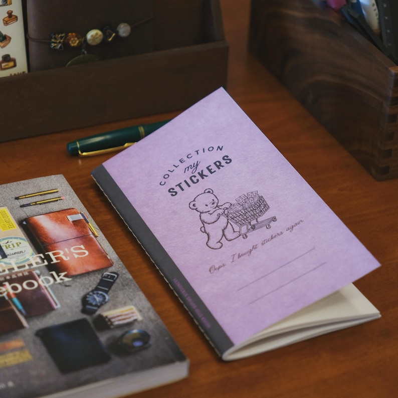 May include: A light purple sticker collection notebook featuring a black line drawing of a bear pushing a shopping cart filled with stickers. The cover includes the text "COLLECTION my STICKERS" and "Oops I bought stickers again" with blank lines.