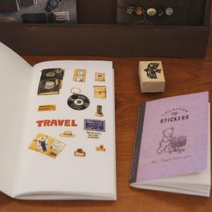 May include: An open notebook featuring watercolor illustrations of travel-themed items including a journal, a vinyl record, a hat, and the word TRAVEL in red. Beside it sits a wooden bird stamp and a purple booklet titled COLLECTION my STICKERS.