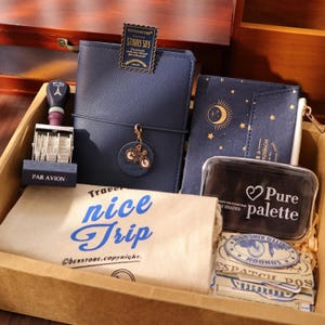 Può includere: Una collezione di articoli di cancelleria a tema viaggio in una scatola di legno. Include un diario blu navy, un timbro, una pochette con un design a luna e stelle e una borsa di tela con la scritta "nice Trip".