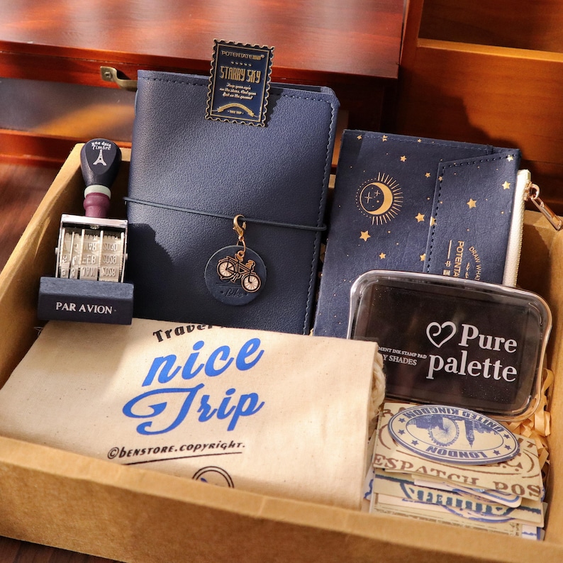 Op de afbeelding: Een verzameling reisbenodigdheden in een houten kist. Inclusief een marineblauw notitieboek, een stempel, een etui met een maan- en sterrenpatroon en een canvas tas met de tekst "nice Trip".