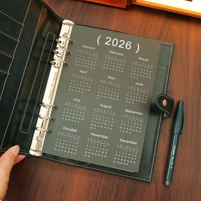 Pu&ograve; includere: Raccoglitore in pelle nera aperto con un inserto calendario 2026. Il calendario &egrave; stampato in bianco su sfondo verde scuro. Una penna nera con inchiostro verde &egrave; appoggiata sulla superficie di legno accanto al raccoglitore.