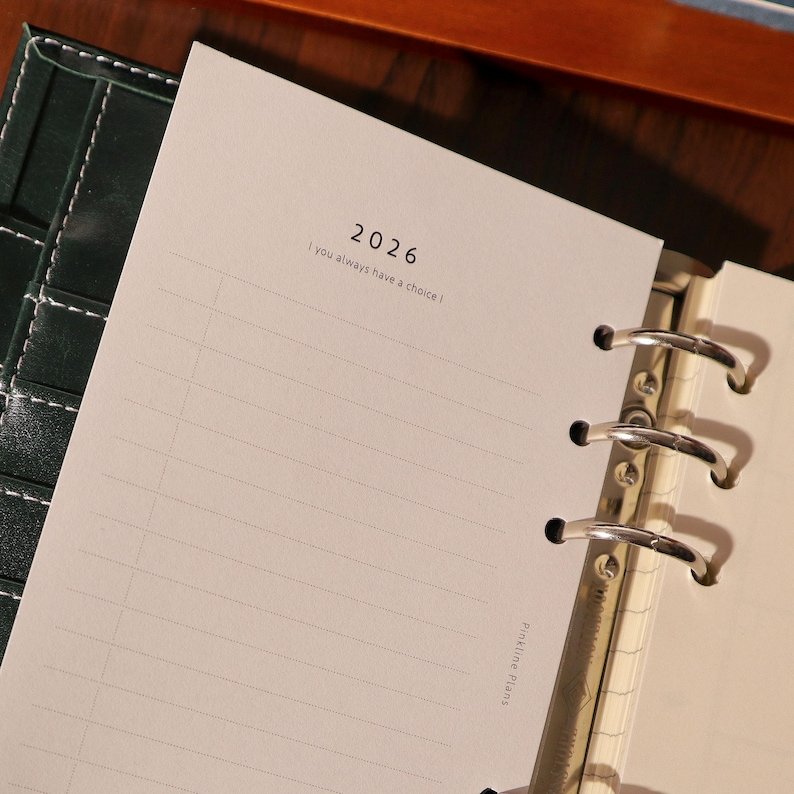 May include: Open planner with lined pages, featuring the year "2026" and the quote "I you always have a choice." The planner has a dark green cover and silver binder rings. The words "Pinkline Plans" are printed on the side of the page.